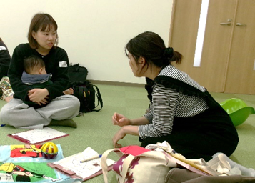 2人の女性の片方が膝に赤ちゃんを抱えながらお互いに床に座って話をしている写真