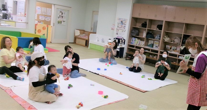 6月ふわふわベビーで絵本を読む保育士とそれを見る親子の様子