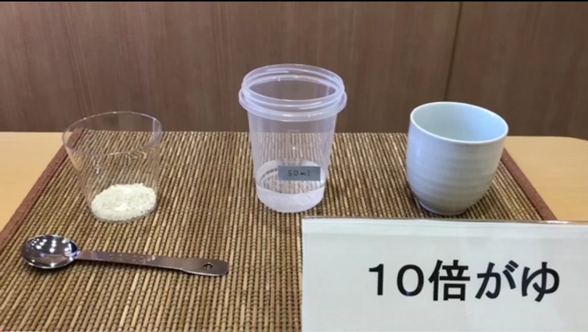 10倍がゆと書かれた立て札のそばにお米が少量入ったカップ、水が入ったプラスチック容器、お椀が並べられている写真（YouTubeへリンク）