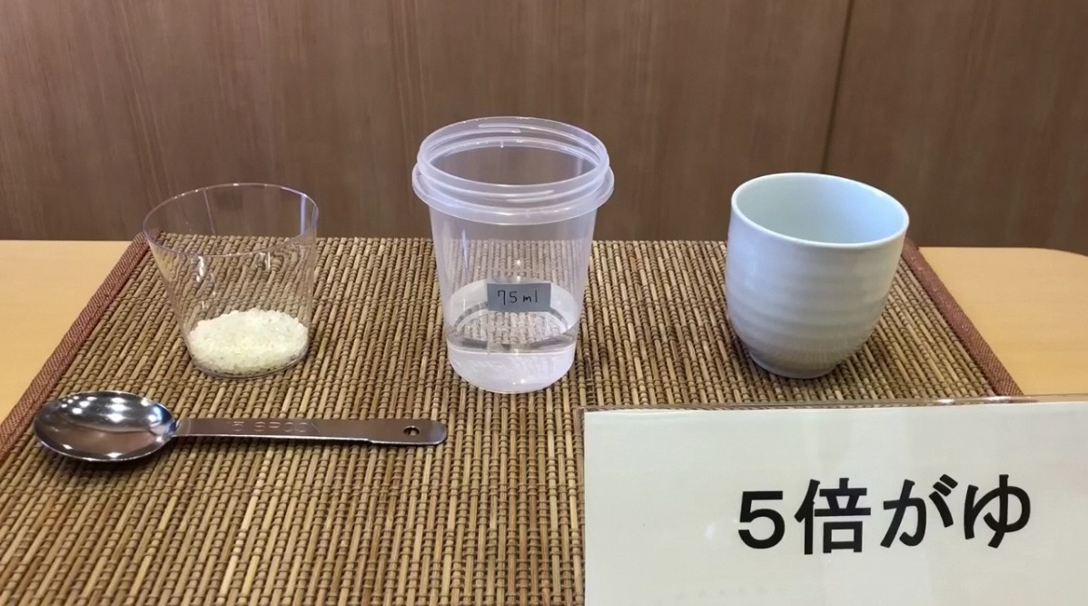 5倍がゆと書かれた立て札のそばにお米が少量入ったカップ、水が入ったプラスチック容器、お椀が並べられている写真（YouTubeへリンク）