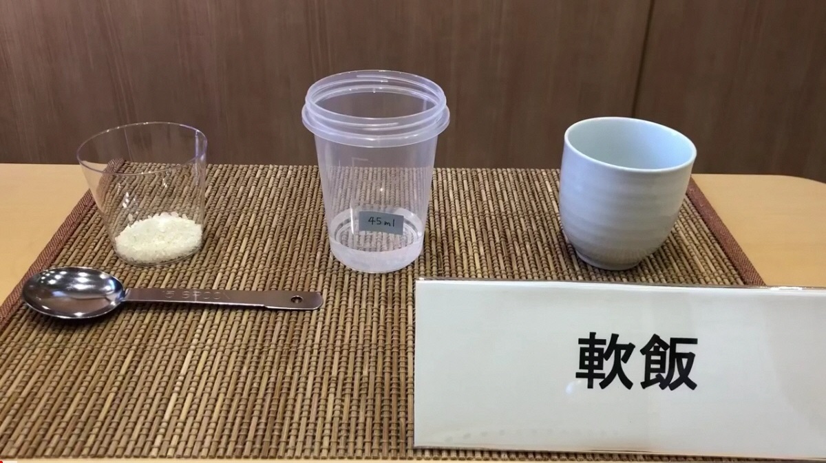 軟飯と書かれた立て札のそばにお米が少量入ったカップ、水が入ったプラスチック容器、お椀が並べられている写真（YouTubeへリンク）
