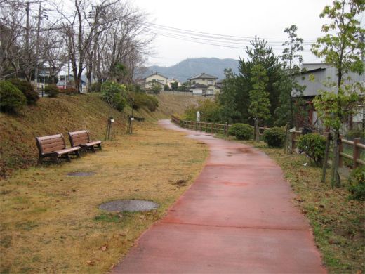 赤色の遊歩道の脇にベンチがある石神緑地の写真