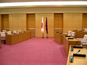 日本国旗と紫の旗の両サイドに議員席が並んでいる議場の写真