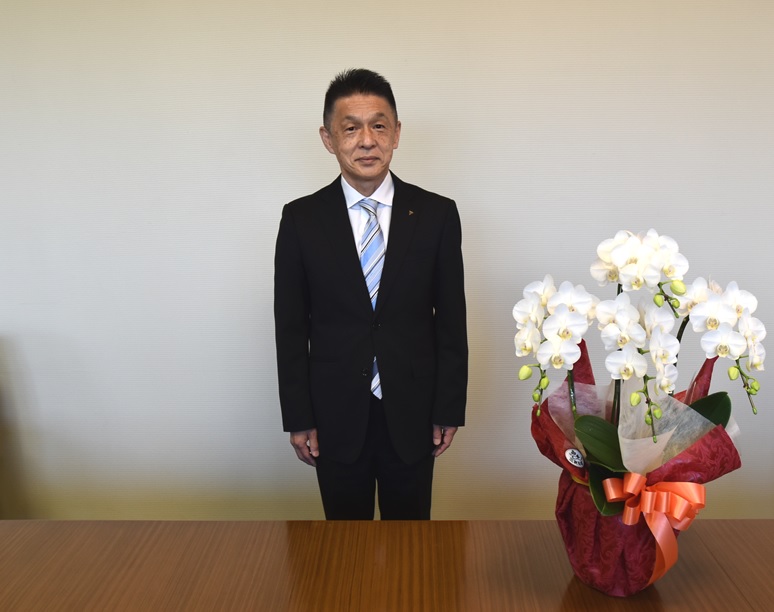 白い花の前で黒いスーツに水色と白のネクタイを付けて立っている平岡弘資氏の写真