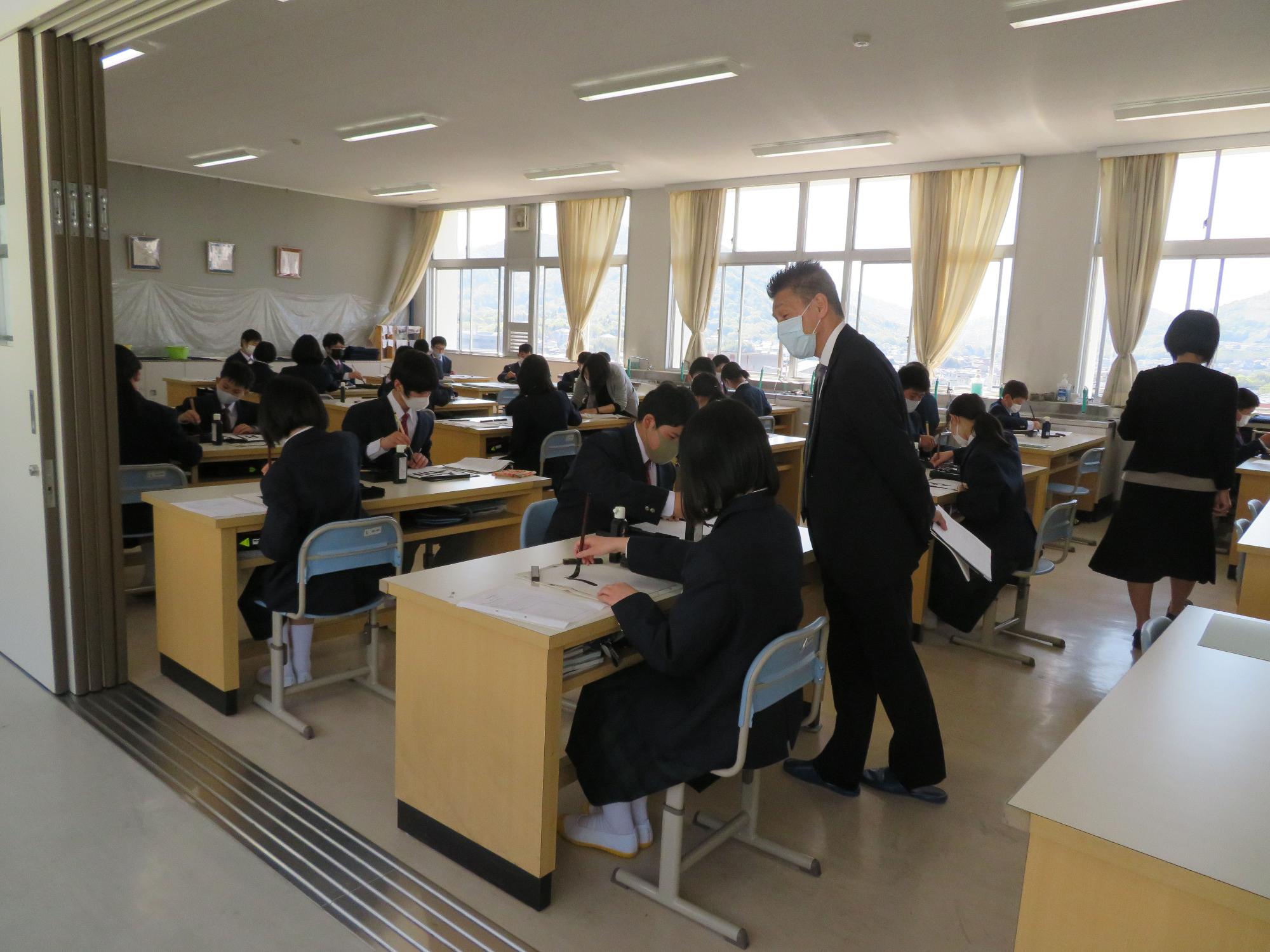 教室で生徒たちが自分の席に座って習字を書いている様子を見学するスーツ姿の男性の写真