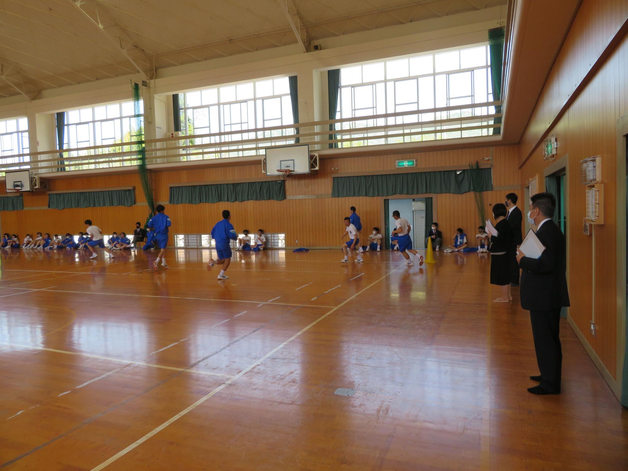 体育館で青い体操着姿の生徒たちがバスケットボールの授業を行っている様子を見学するスーツ姿の男性の写真