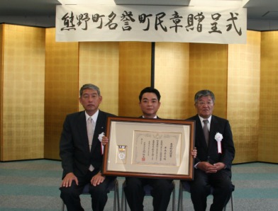 室内でスーツを着た男性3名が椅子に座り額縁に入れた賞状を持っている写真