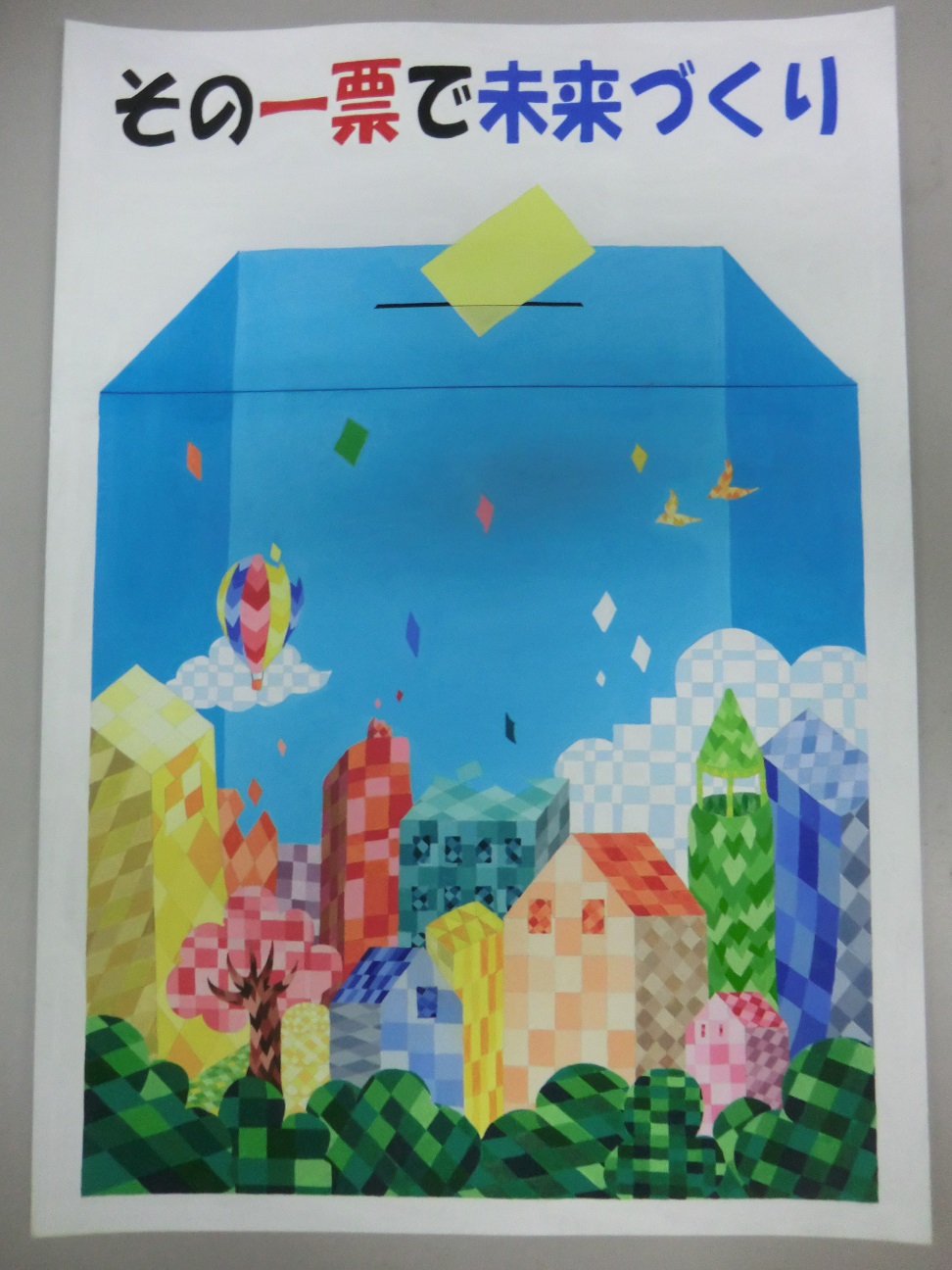 「その一票で未来づくり」カラフルな町が描かれた投票箱のポスターの写真