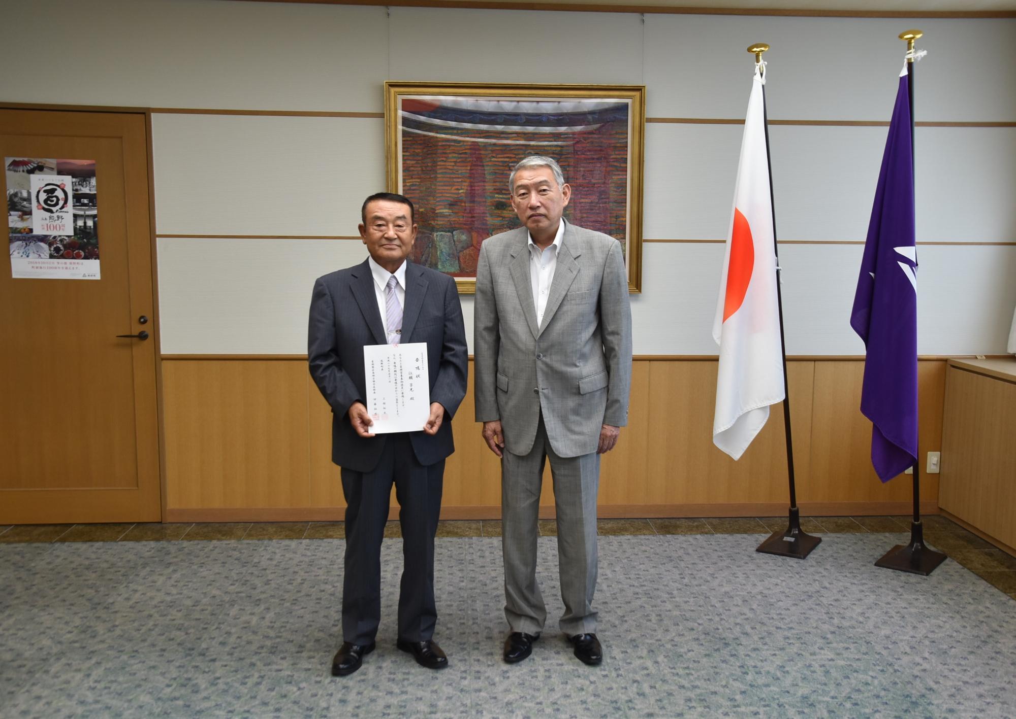 室内でスーツを着た男性2人が正面を向いて委託式を行っている写真