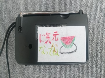 手書きのスイカが描かれた紙に黒色のラジオにテープで止められている写真