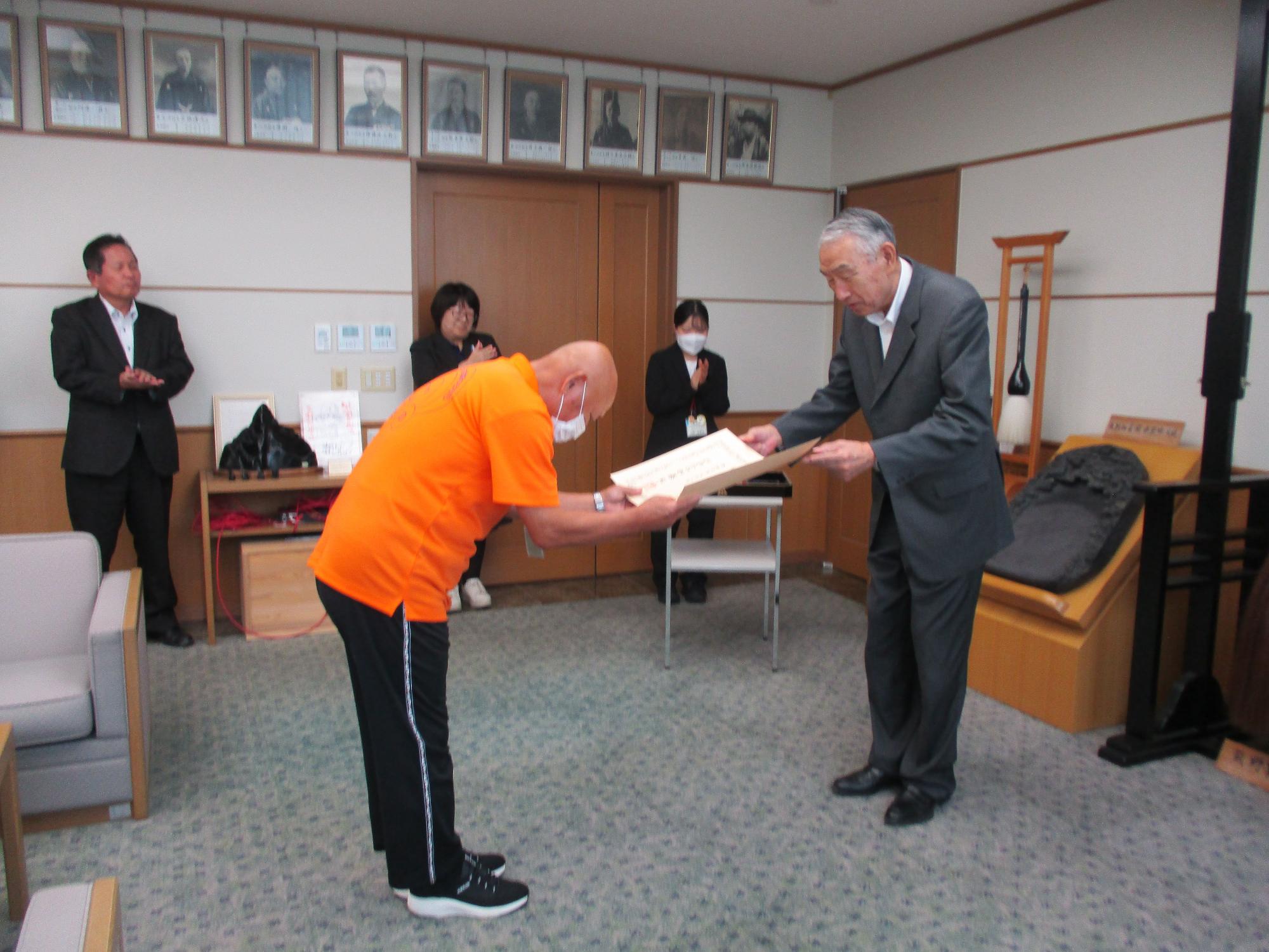 伝達式で熊野町長から書状を受け取るオレンジ色のシャツを着た男性の写真