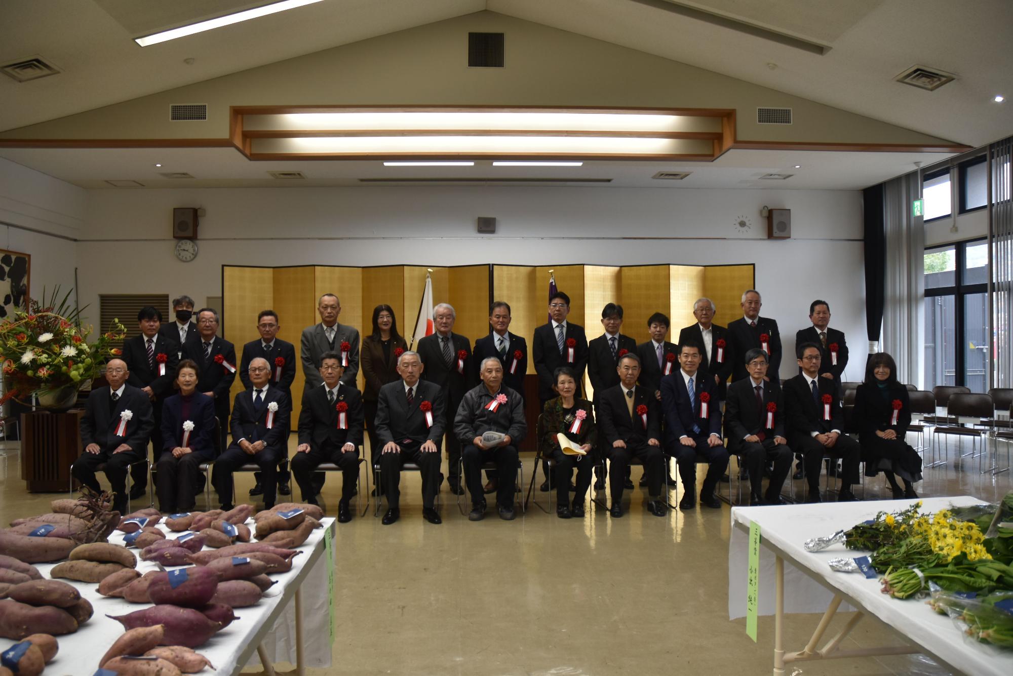 テーブルに並べられたさつまいもやほうれん草などの農作物といっしょに記念撮影に臨む熊野町長や関係者の方々の写真