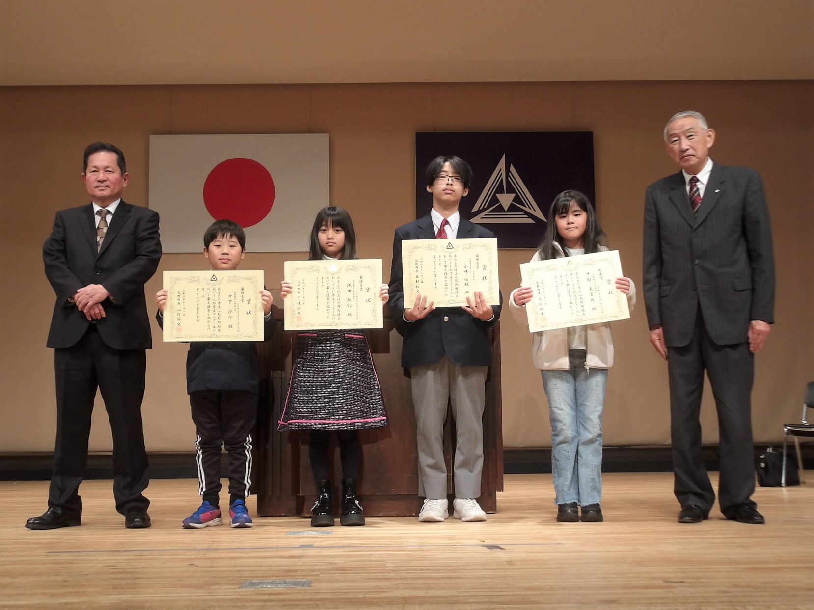 表彰式の壇上で熊野町長といっしょに表彰状を持って記念撮影に臨む四名の児童と生徒たちの写真