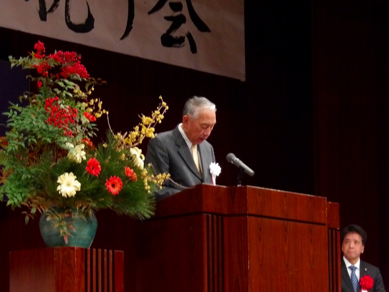 花が飾られた演台でお祝いの言葉を述べる熊野町長の写真