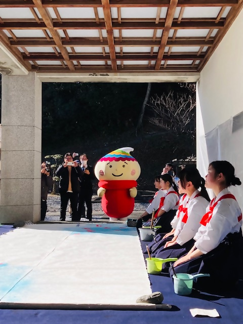 ゆるキャラ「ふでりん」が見守る中、巨大な半紙の前で正座している女子生徒たちの写真