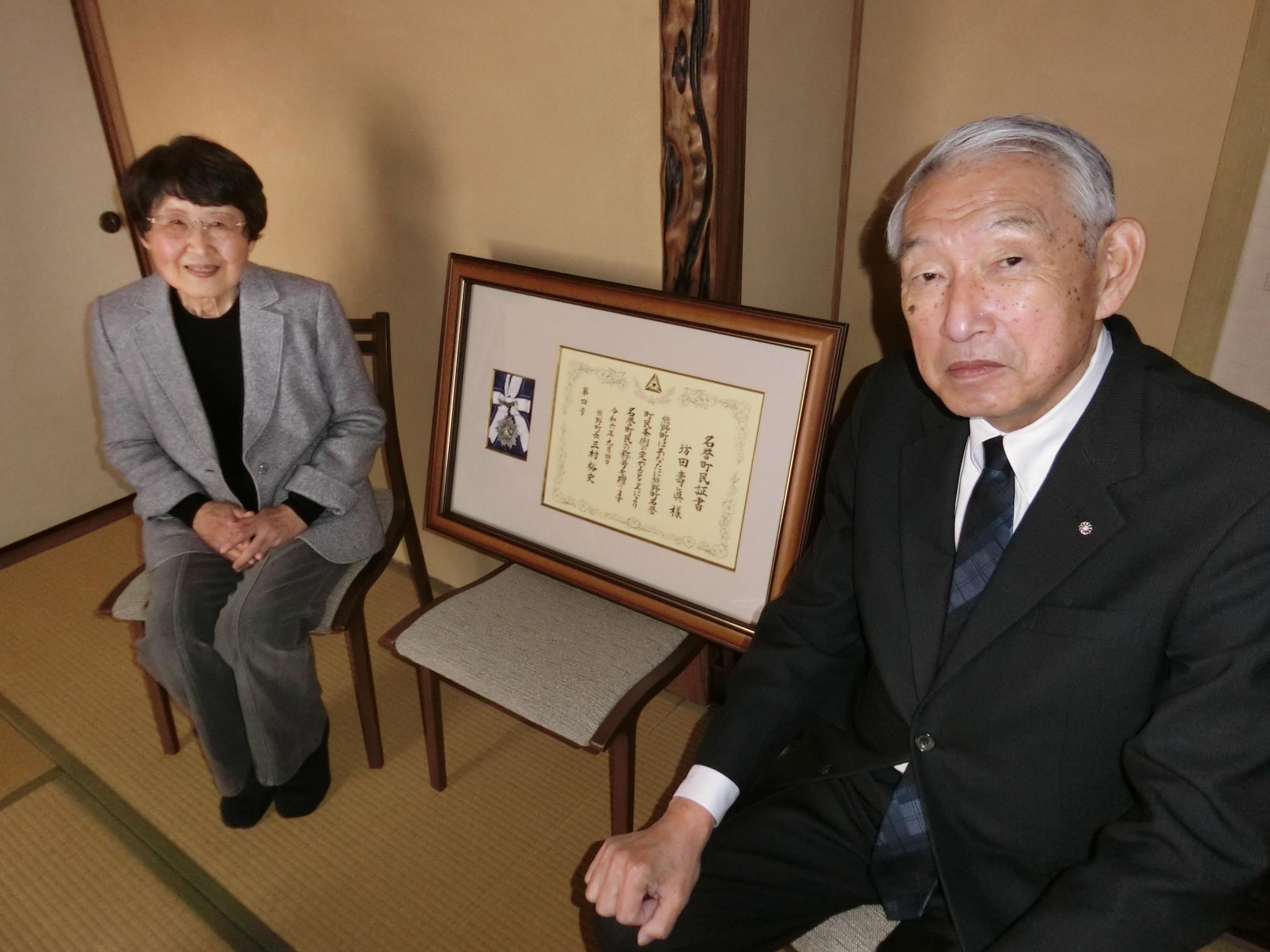 名誉町民の表彰状を挟んで椅子に座り記念撮影に臨む熊野町長と真砂久子さんの写真
