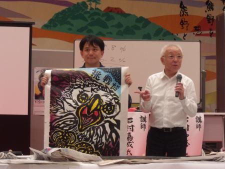 「挑戦だ」と描かれたドローイング作品を手にしている石村嘉成さんと作品の解説をしている父親の和徳さんの写真