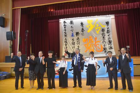 講堂で舞台に飾られた席書をバックに記念撮影に臨む筆まつりの参加者たちの写真