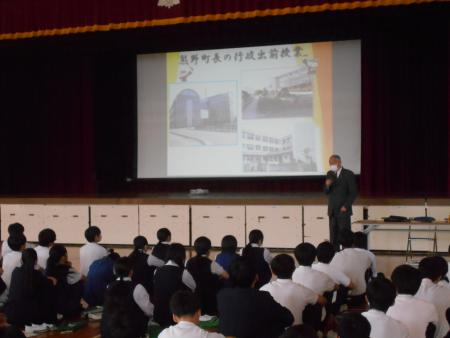 舞台上に大きなスクリーンがありその前で話す男性の写真