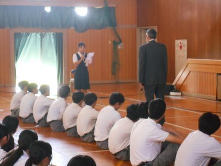 体育館で座っている生徒たちの前で話す女子生徒と対峙している男性の写真