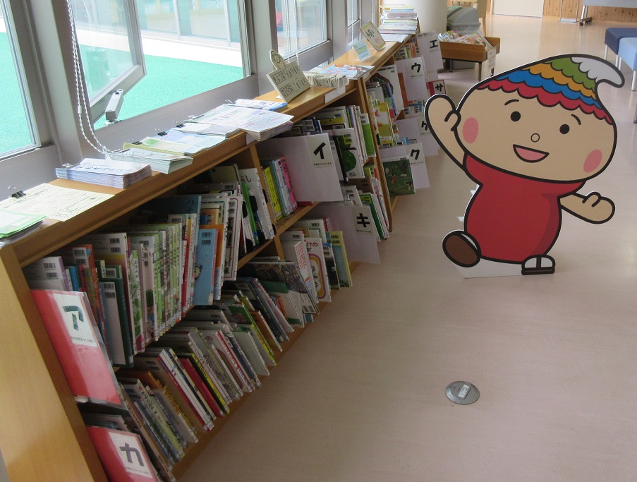 窓に沿って並ぶ本棚に本が並べられており、そのそばにふでりんの立て看板がある写真