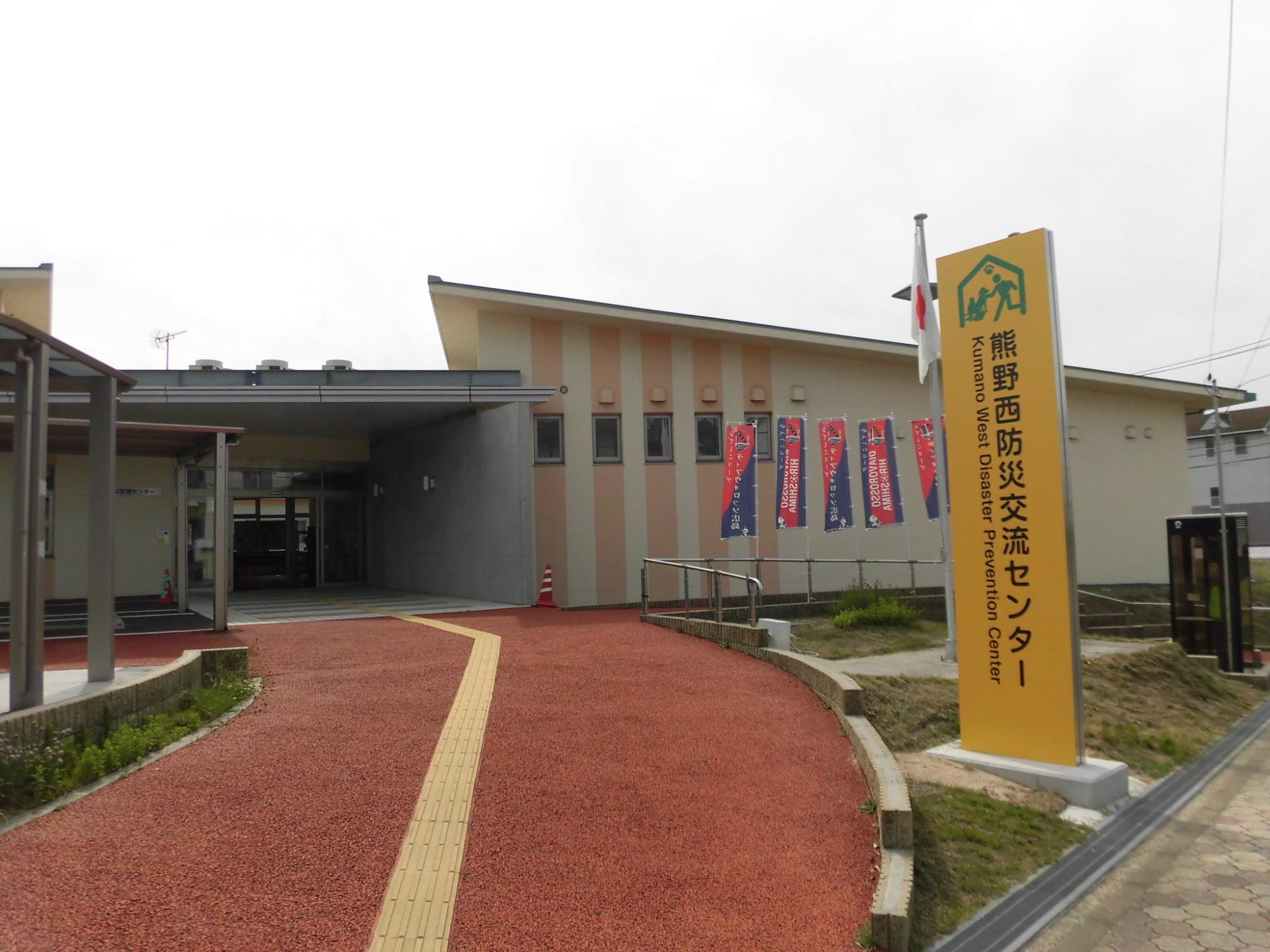 橙色の看板と点字ブロックの設置された赤い歩道の奥に熊野西防災交流センターの建物の写真