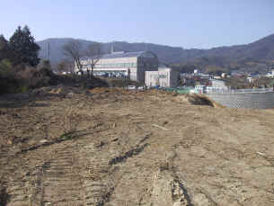 平らな土の地面になっている計画地の写真。役場庁舎の丸い屋根が確認できる。