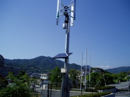 柱の上部に風力発電の風車があり、中ほどの高さに太陽光発電のパネルが設置されている発電設備の写真