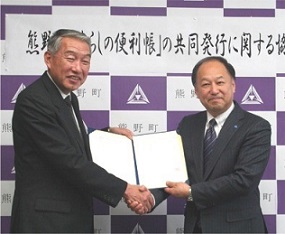 協定書を掲げて握手をする株式会社サイネックスの代表者と三村町長の写真