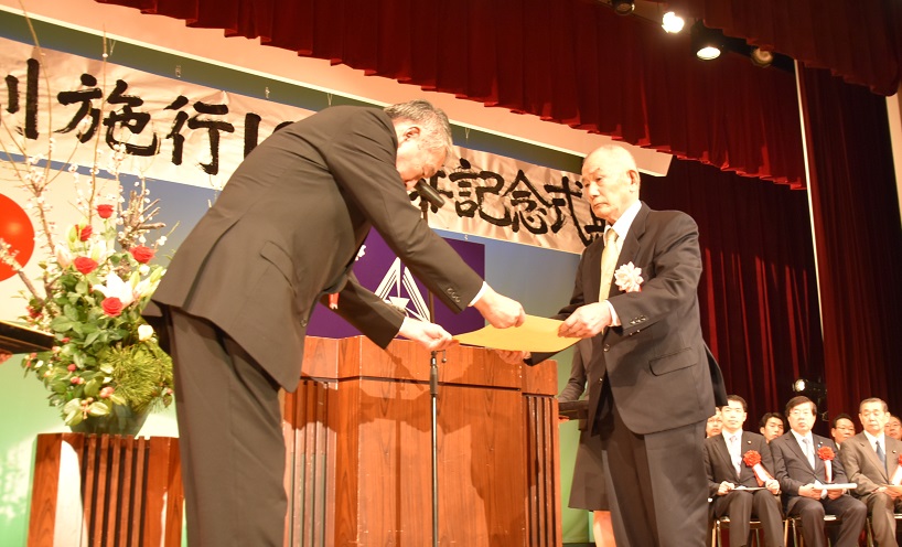 表彰式にて、授与された表彰状を受け取る男性の写真