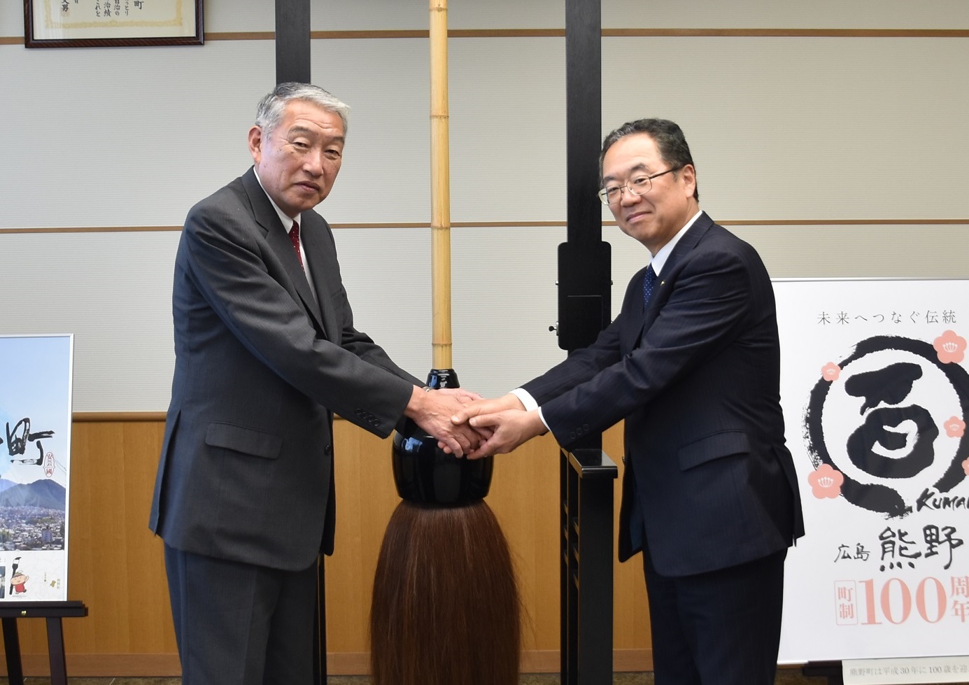 熊野筆の前で笑顔を浮かべ握手を交わす川上市長と三村町長の写真