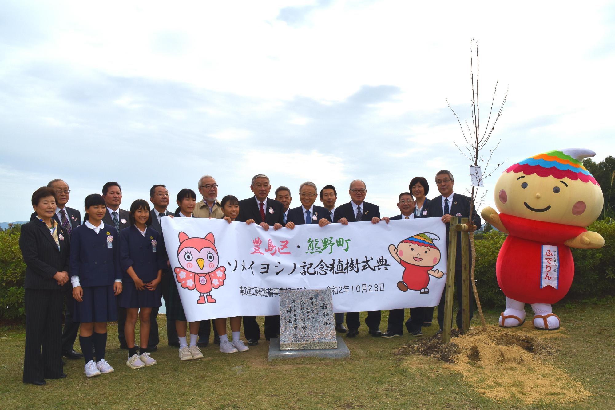 豊島区と熊野町のソメイヨシノ記念植樹式典にて、手に横断幕を掲げ石碑の後ろに並ぶ参加者たちとふでりんの写真