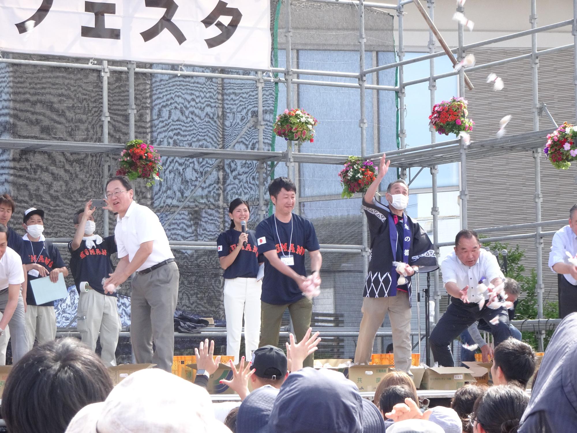 屋外の仮設ステージの上から数人の男女が餅を投げている「もちほり」の写真