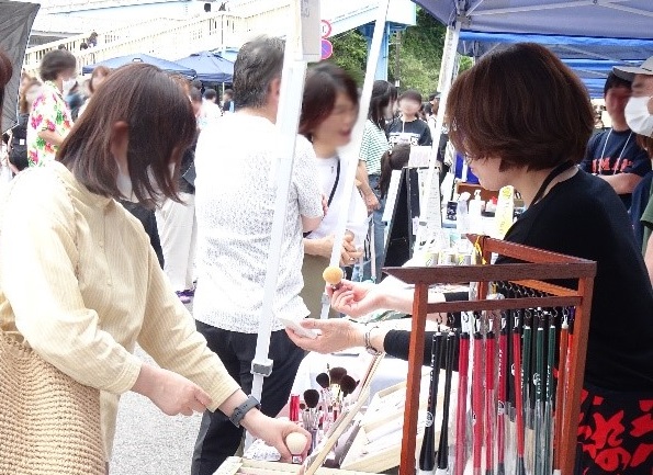 「オール熊野マルシェ」でスタッフから説明を受けながら筆を手に取る女性客の写真
