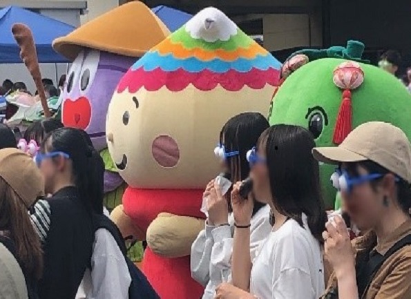 屋外で個性豊かなご当地キャラクターと青い眼鏡をかけた人々が並んでいる写真