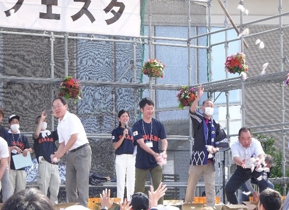屋外の仮設ステージの上から数人の男女が笑顔で餅を投げている写真