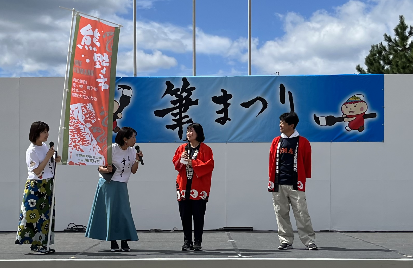 ふれあいステージの壇上で、白いシャツを着た司会者が赤い法被を着た熊野市職員に質問している写真