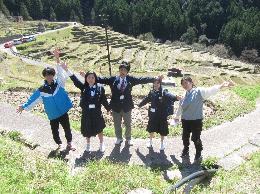 山間に広がる棚田の前で5人の生徒が腕を広げてポーズをとっている写真