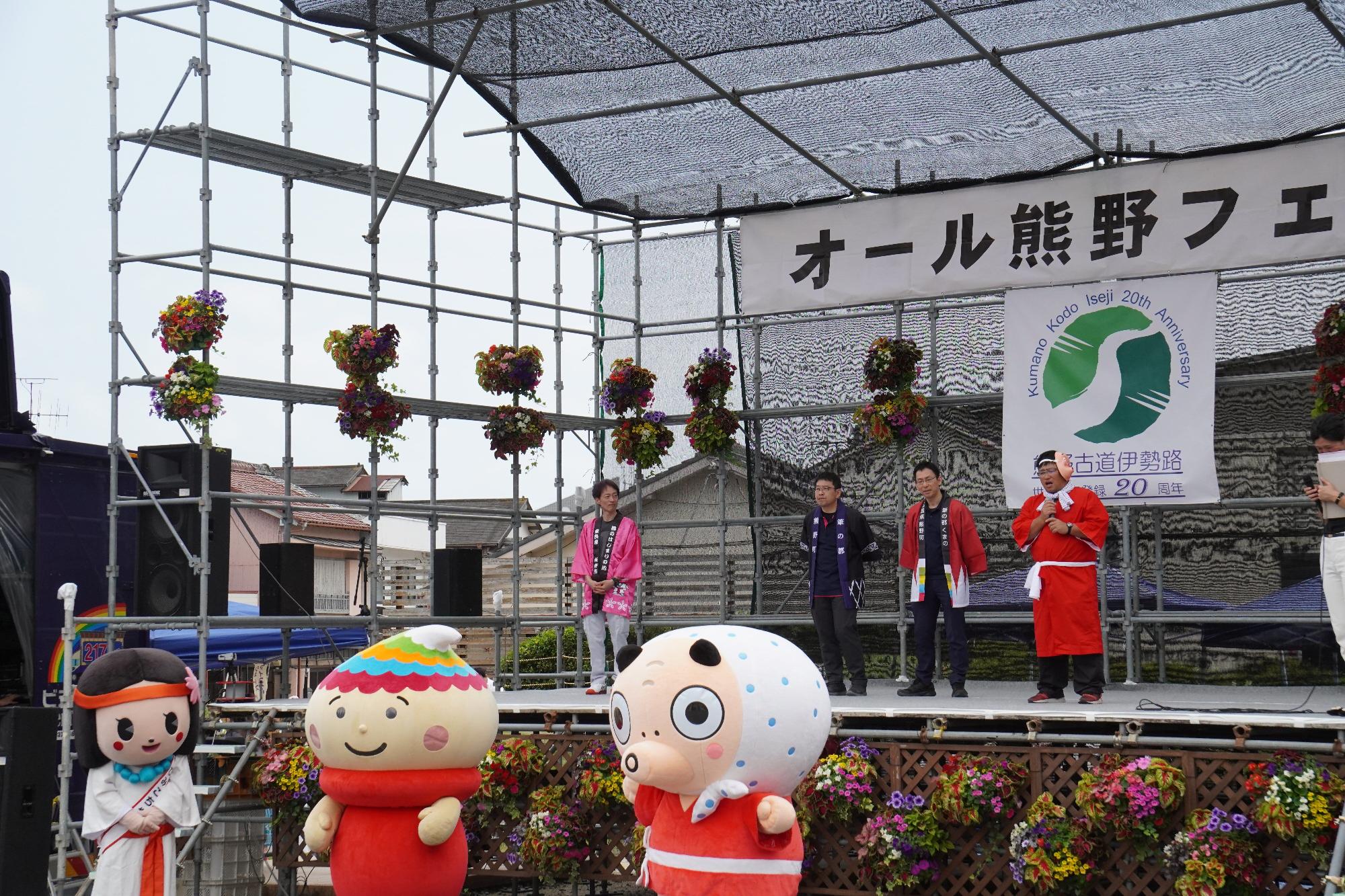 3体のキャラクターがステージ前に並び、ステージ上の代表者が自分たちの町や市について紹介している写真