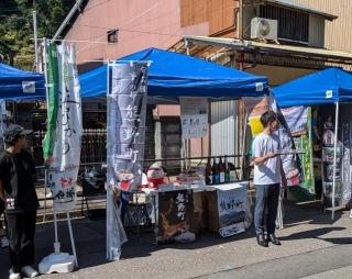「筆の都 熊野町」と書かれた幟が立っている青いテントの下に設置された熊野町特産品販売ブースの写真