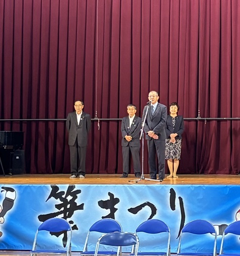 筆まつりの会場で壇上からマイク越しに挨拶されるスーツ姿の男性たちの写真