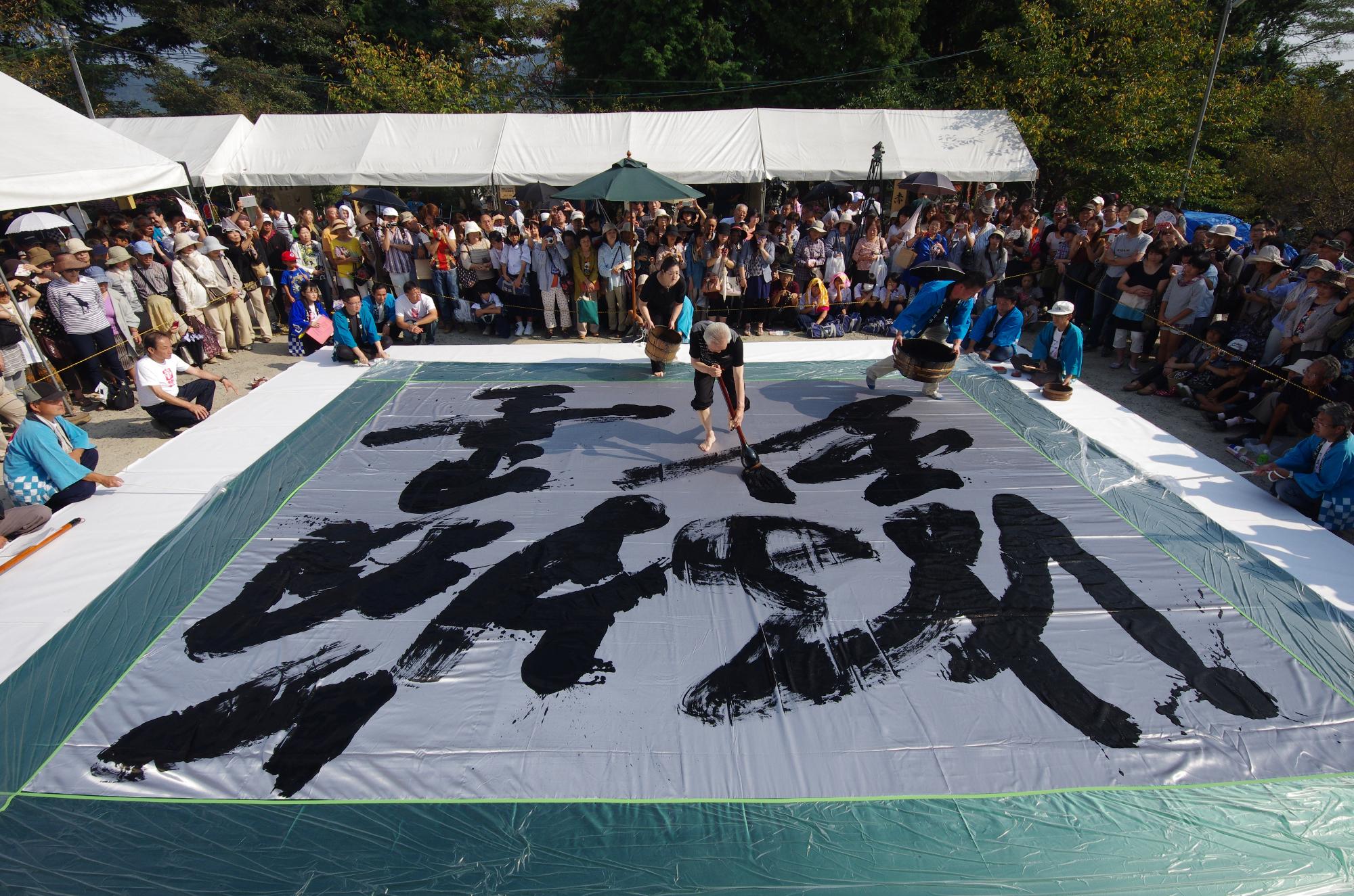 筆まつりの会場で巨大な布に大筆で文字を書いている書道家の写真