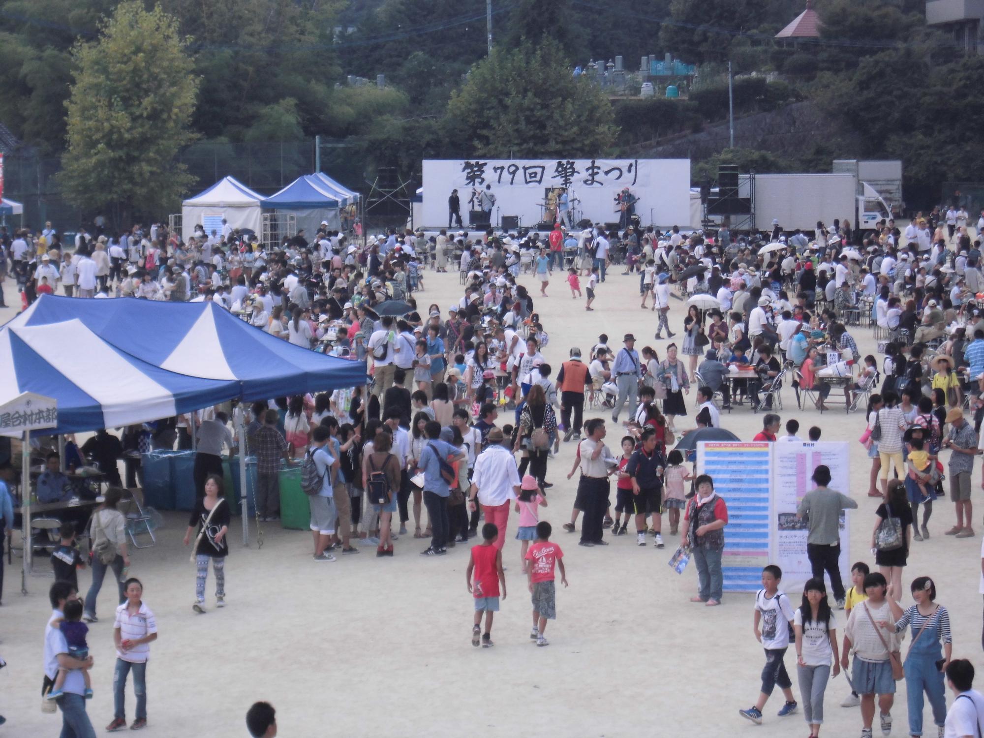 青や白の屋根のテントや野外ステージがあり多くの人々で賑わっている筆まつりの会場の様子を捉えた写真
