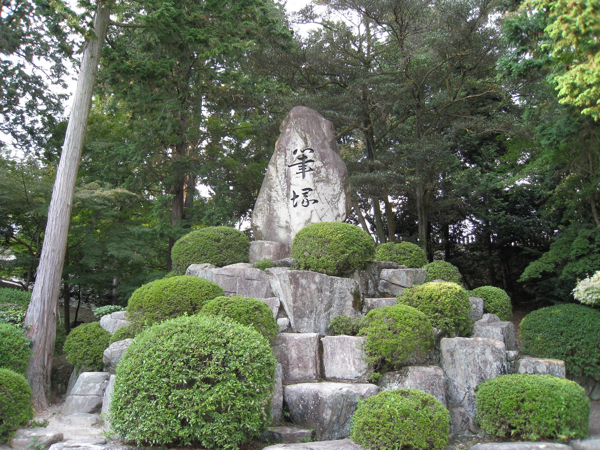 周囲を木々に囲まれた中、数段の石段とその脇に綺麗に整えられた植木があり、その先に「筆塚」と刻まれた石碑がある写真。