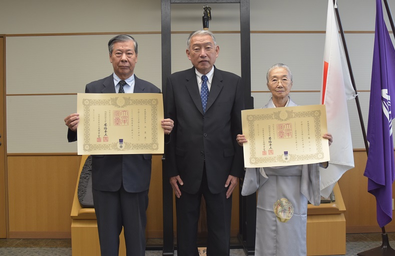 中央に男性が立ち、写真右に着物を着た女性、写真左にスーツ姿の男性が立ち、それぞれが授与された賞状を掲げている写真