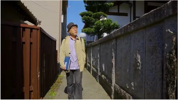 狭い路地をスケッチブックを持って歩く野村重存さんの写真