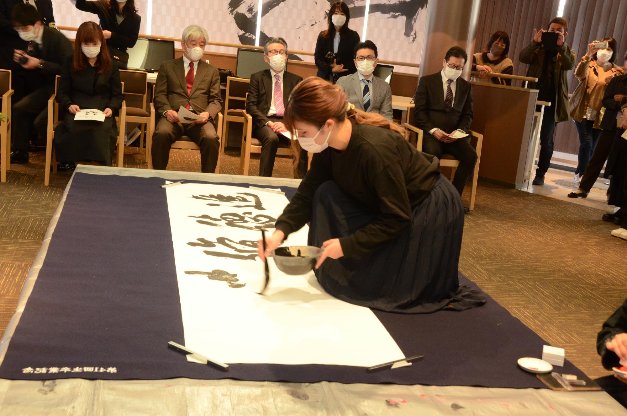 大きな半紙に毛筆で書道を行う黒い服を着た女性の写真