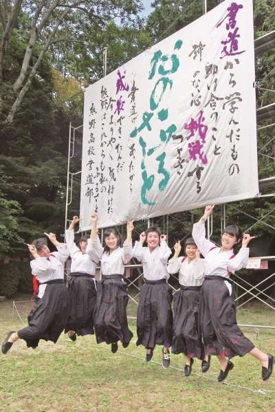背後に掲げられた毛筆で書かれた大きな垂れ幕の前でジャンプをしている人々の写真