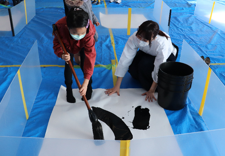 ブルーシートが敷かれた広間で大筆を使って書道を行う二人の女性の写真