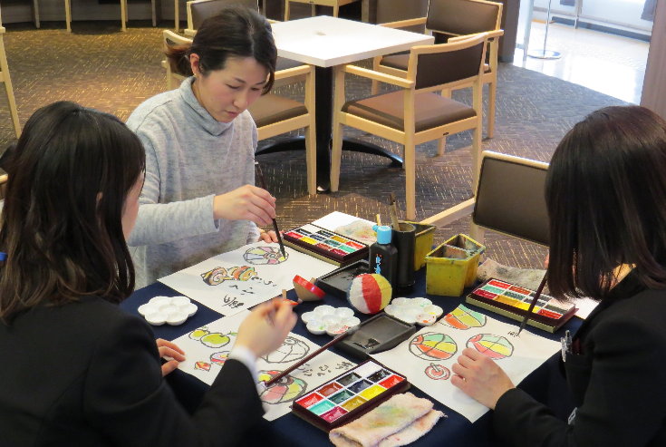 工房の教室内で筆を使った絵画を作っている三名の女性の写真